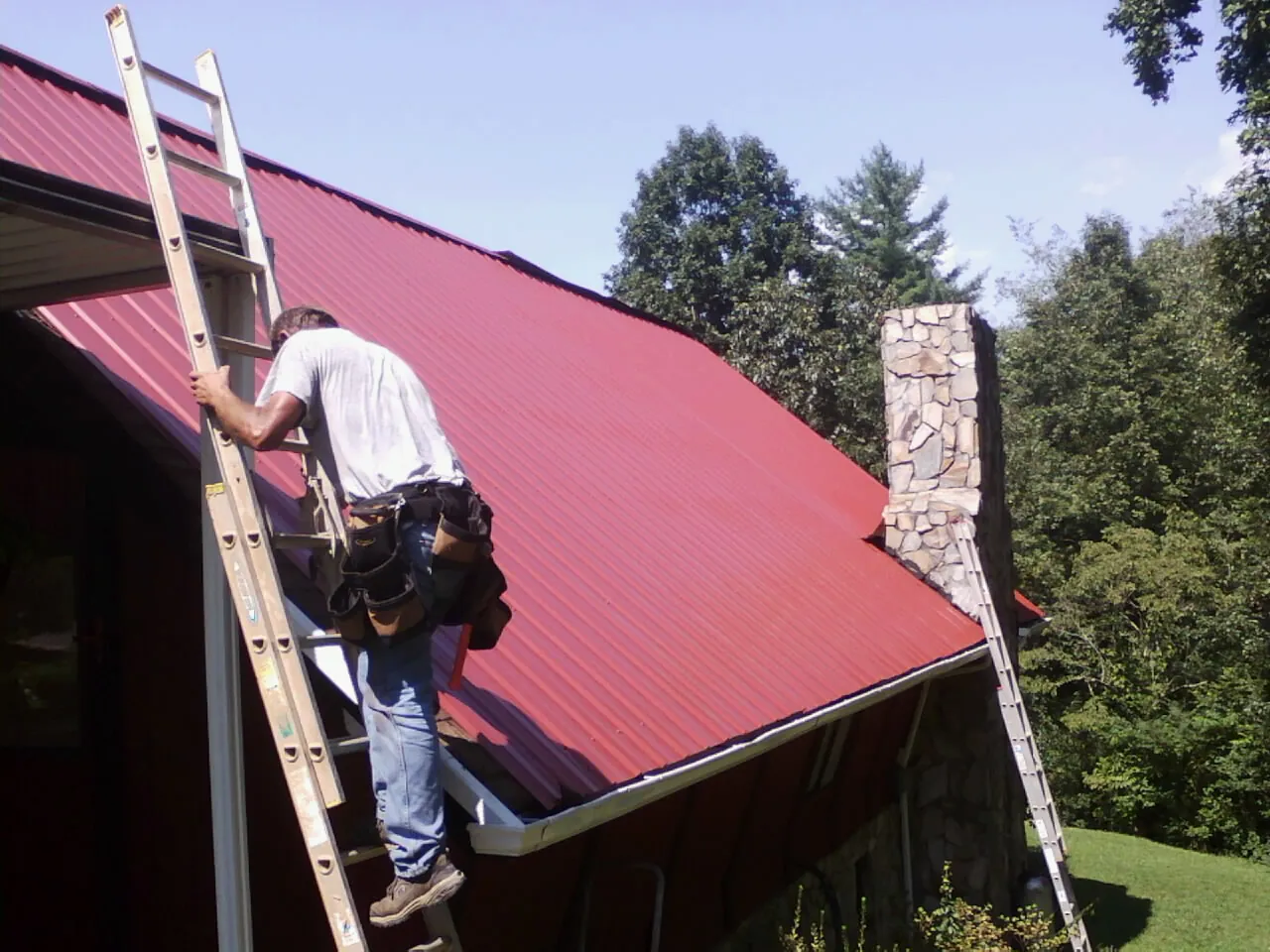 Roofer on a ladder beside a red metal roof and stone chimney — Soffit & Fascia Repair by Roofing Brothers in Gibsonton